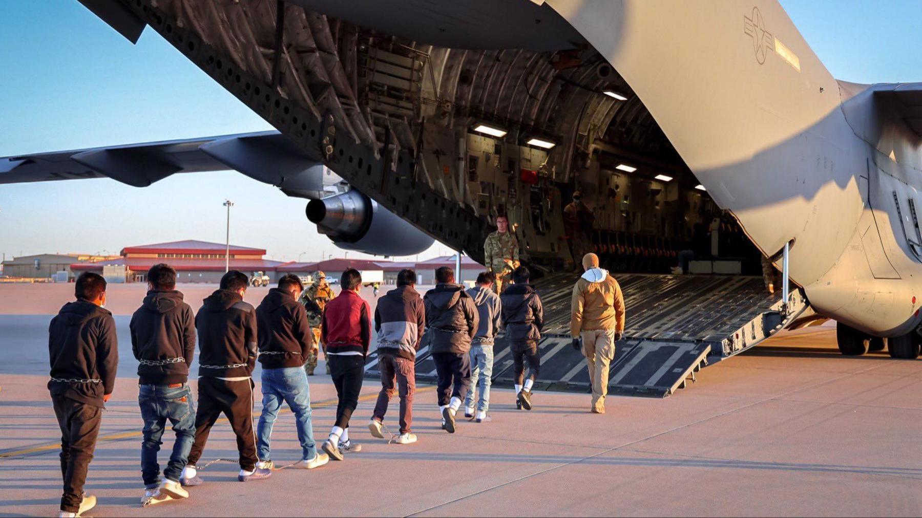 A line of men, wearing chains around their waists, walk with their backs to the camera onto a US military plane 