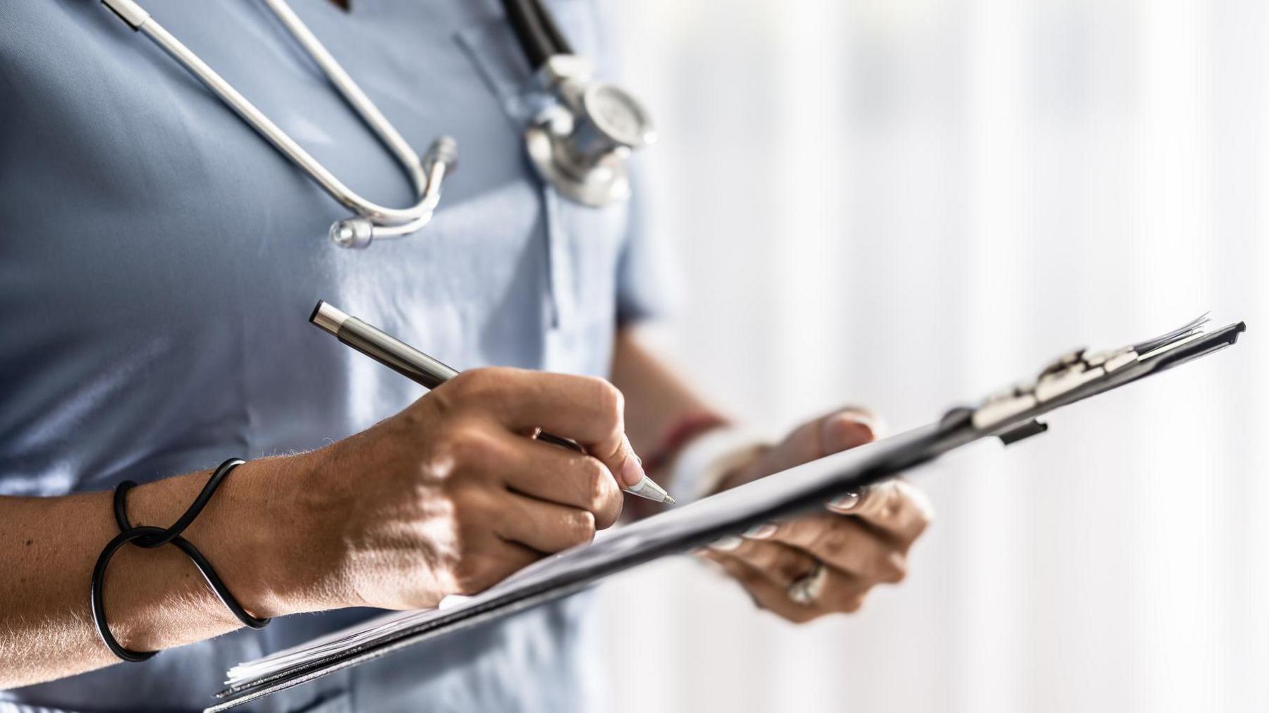 A health professional wearing a blue uniform is using a pen to write on a sheet on a clipboard. They have a silver and black stethoscope around their neck and a black bracelet on their wrist. They are wearing a ring on their left hand. The background is blurred.