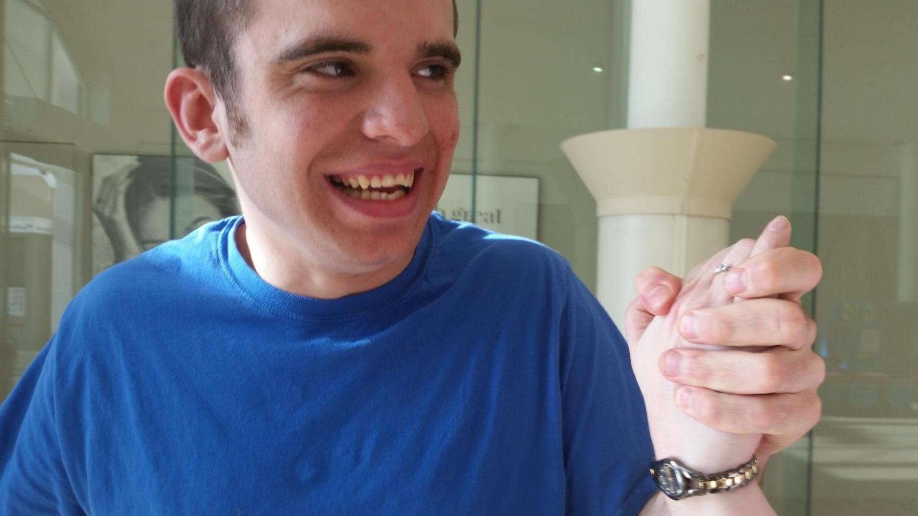 Declan Morrison holding the hand of someone who is not in shot. He is wearing a royal blue T-shirt and is smiling animatedly at the person holding his hand to the right of shot. He has dark brown hair and dark eyes.