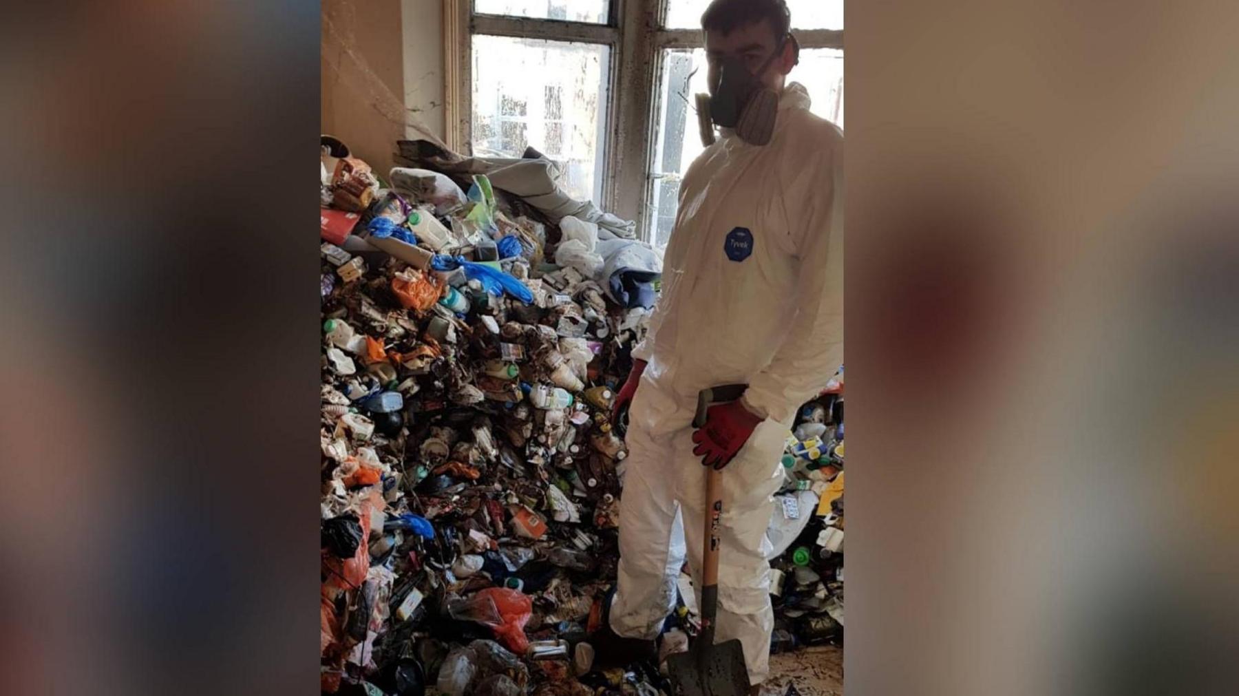 Photograph shows a picture of a pile of rubbish mounted up in the bay of a window. A boy stands in front of the pile of rubbish. He wears full PPE, a face mask and holds a shovel. He wears black and red gloves.