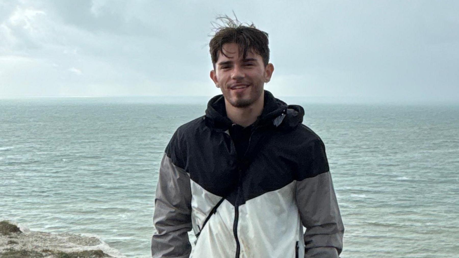 Oliver Archer in a grey, black and white patterned coat, standing in front of the sea. He has dark hair and a beard.