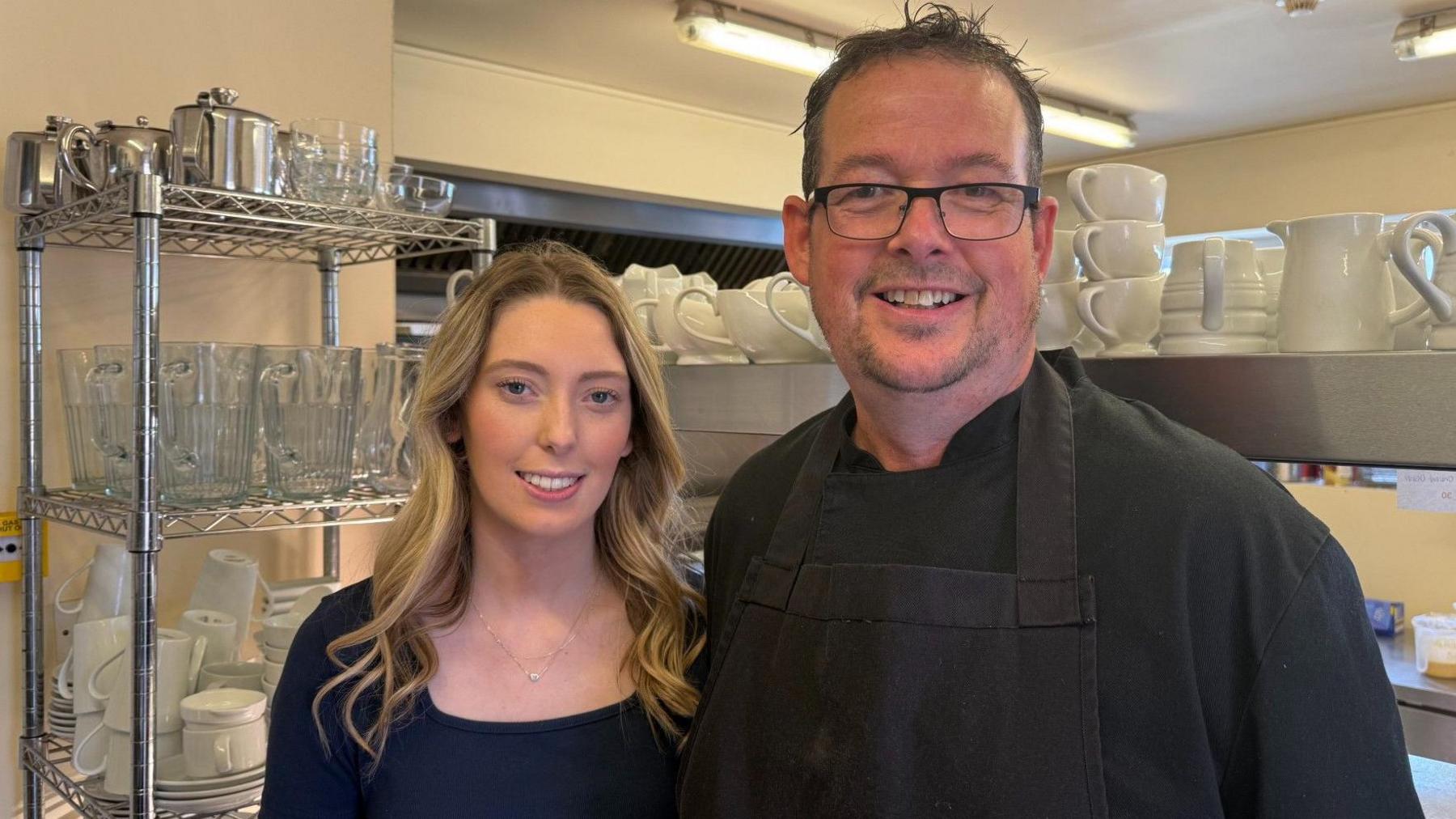 A young woman with long hair standing next to a man with glasses and a goatee beard. He is wearing a black shirt and an apron. There are racks of cups and saucers in the background.
