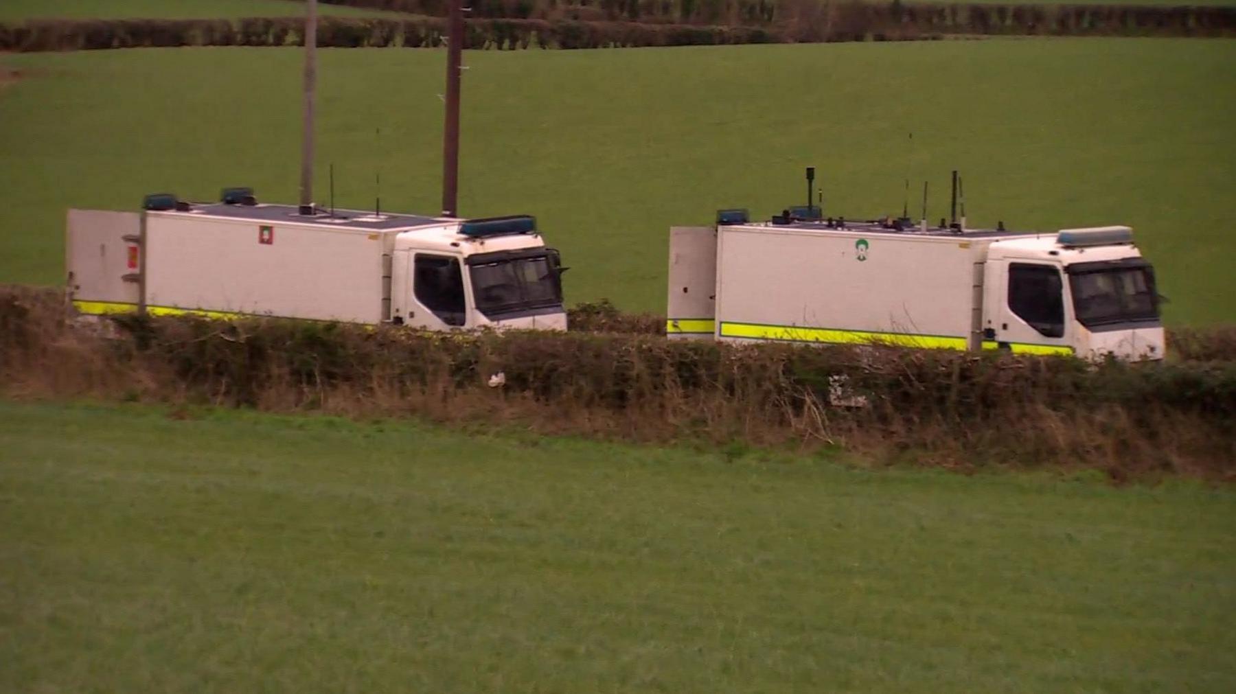 Two white truck, used for bomb disposle, sit infront of a green grassed field.