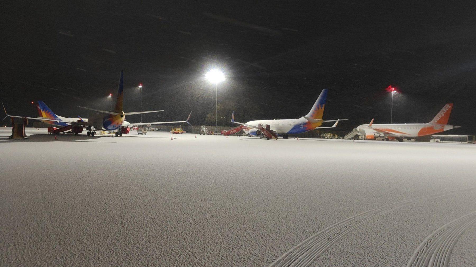 A runway area covered in snow is shown at Bristol Airport, with four passenger planes visible in the background, two of them with Easyjet livery. There is a pair of tyre tracks made by a vehicle in the foreground