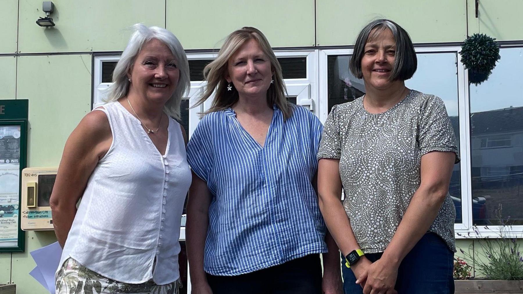 Three middle-aged women stand in front of a pale green building. A sign reading "The Buckland Centre" is on the wall behind them.