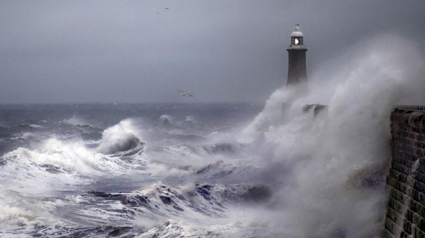 Waves whipped up by a storm crash against a sea wall in Tynemouth, with a lighthouse in the background.