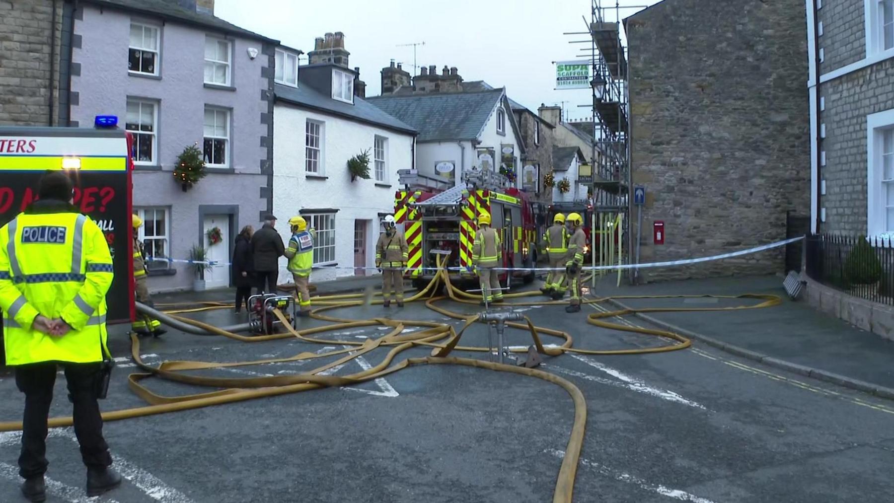 View down a street in which stand firefighters and two fire engines, with a tangle of fire hoses on the ground. A police officer stands on the left of the scene and a firefighter is talking to a man and a woman dressed in dark clothing.