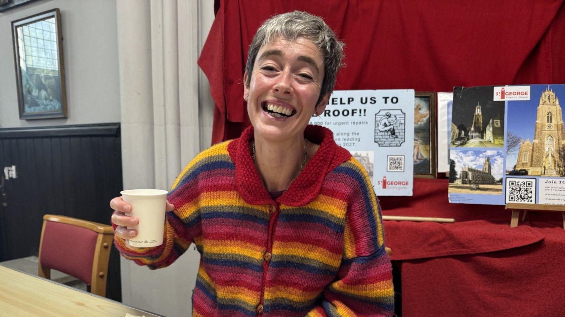 A woman in a multi-coloured stripy top smiles widely while holding a paper cup in the air.