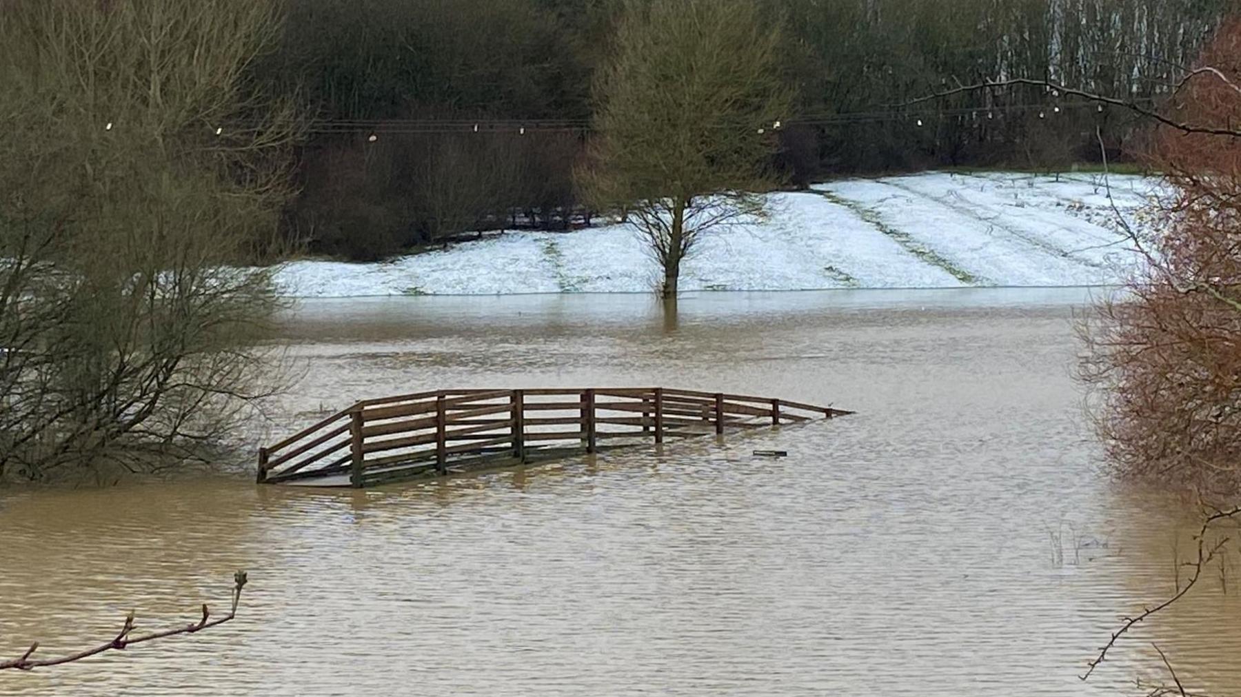 The wooden railings of a bridge half-submerged in brown water. Snow-covered fields can be seen in the background, as well as partially underwater trees.