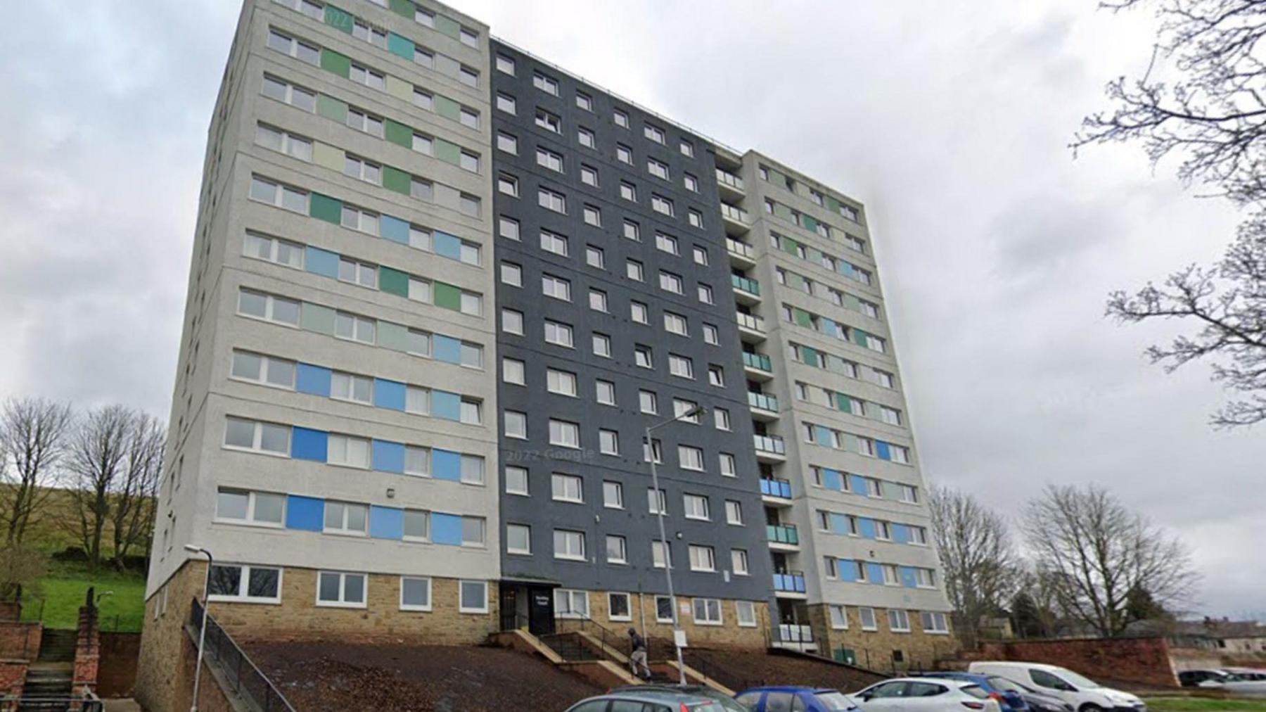 An apartment building stands against a grey sky with cars parked in front.