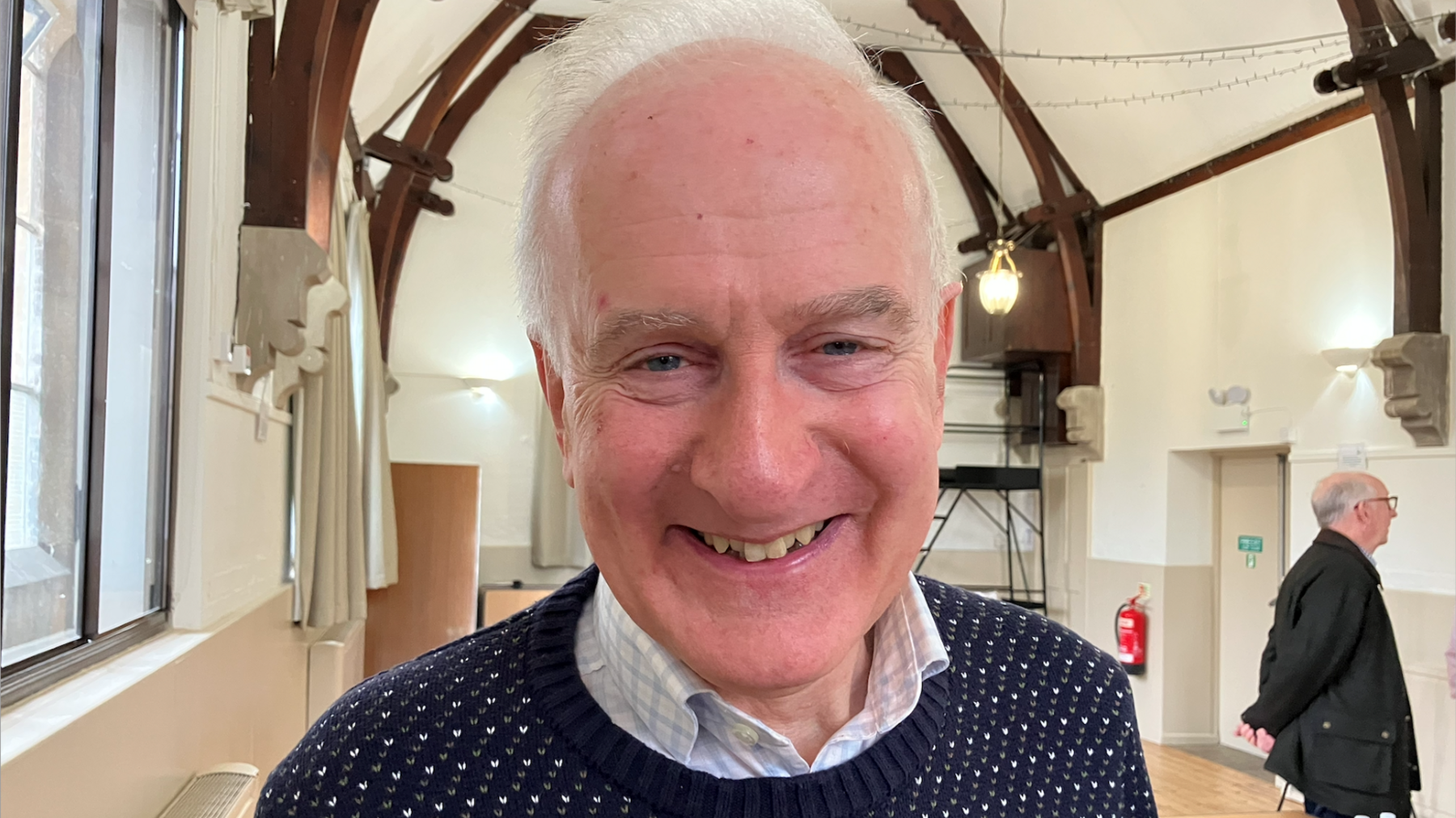 Richard Irvin, standing in a room, with wooden beams and white walls, with another man to the right. He is smiling and wearing a checked shirt, blue and white patterned jumper, with short grey hair. 