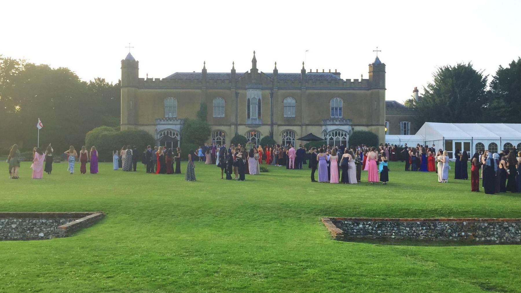 An evening photo head on of the house, with lots of people milling around on the grass in prom dresses and suits. The lawn is very green.
