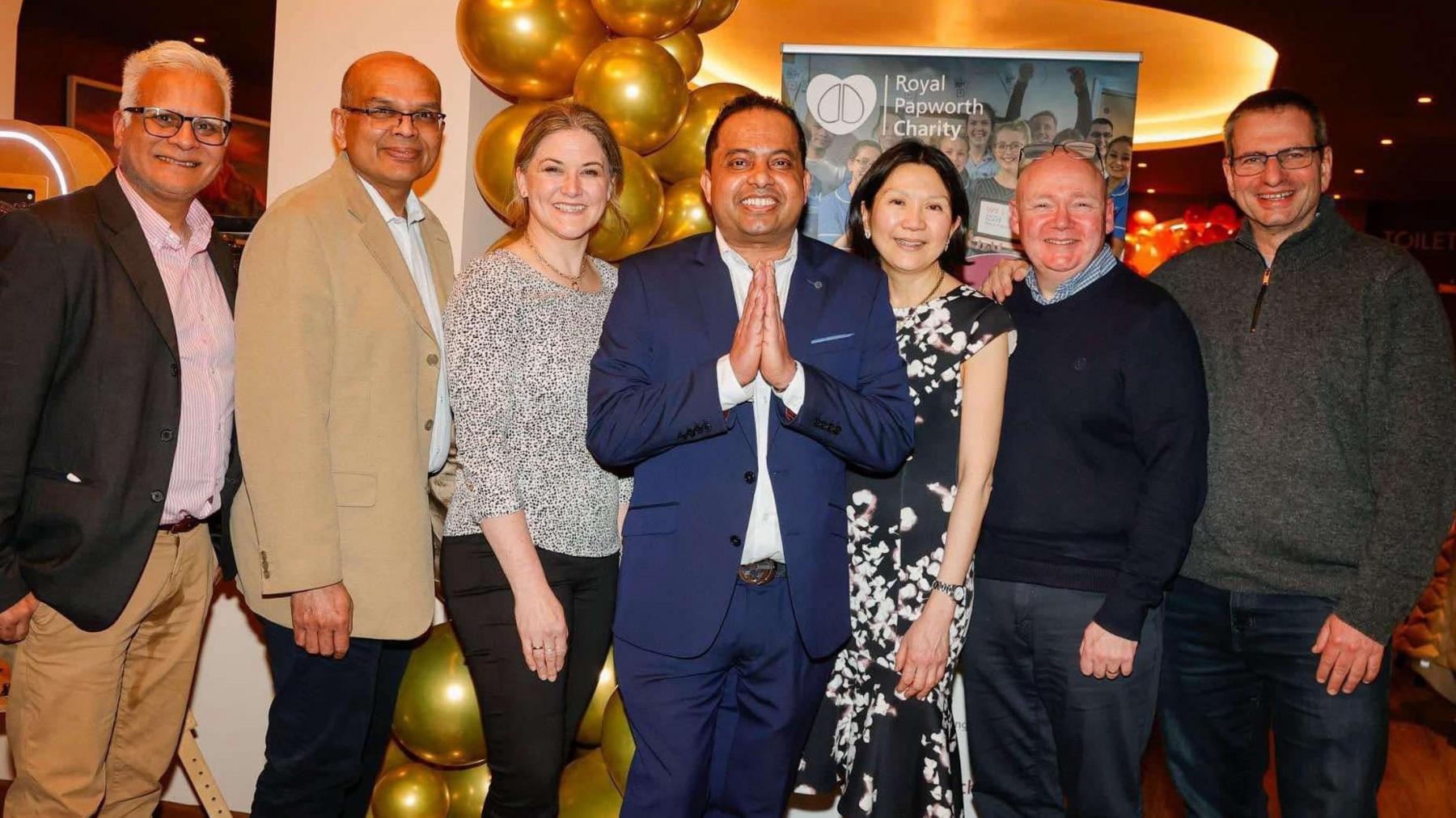 Mr Bashyal stands in the centre of a row of six other people, with his hands in a prayer gesture. The four male and two female guests are standing in front of a gold balloon display and Royal Papworth Charity banner in the restaurant.