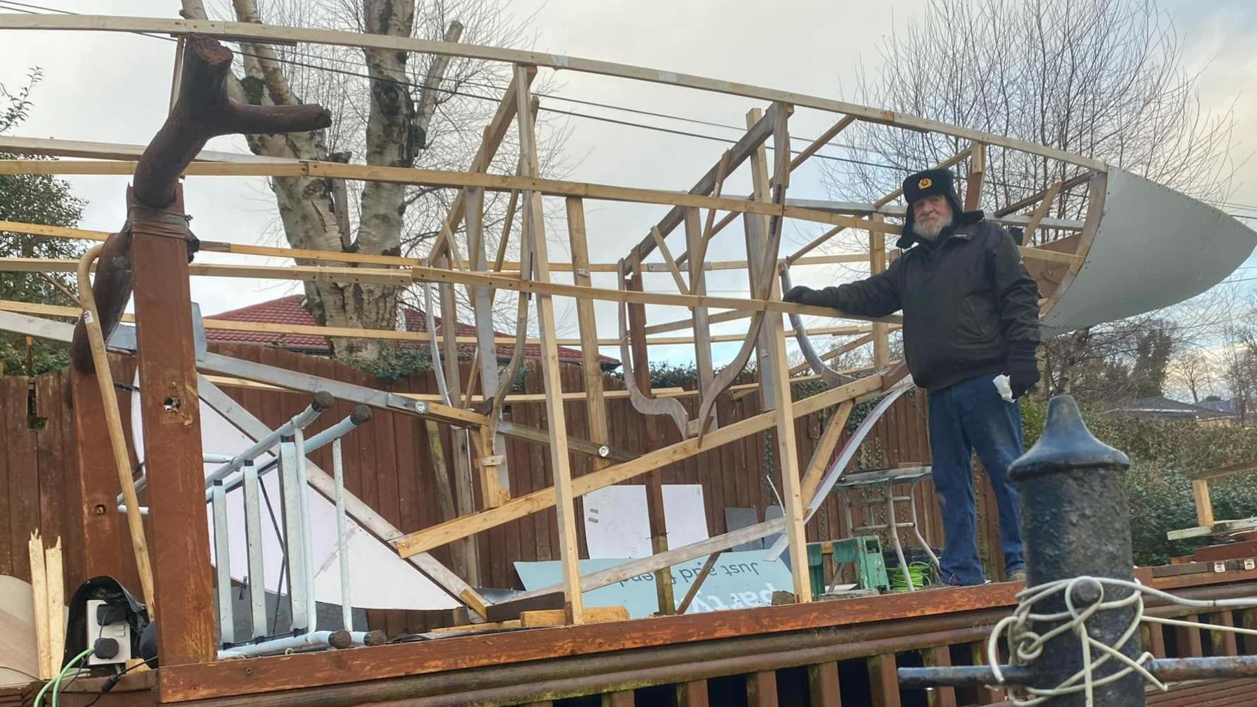 A man, Ian, stands in front of a part-constructed boat. 