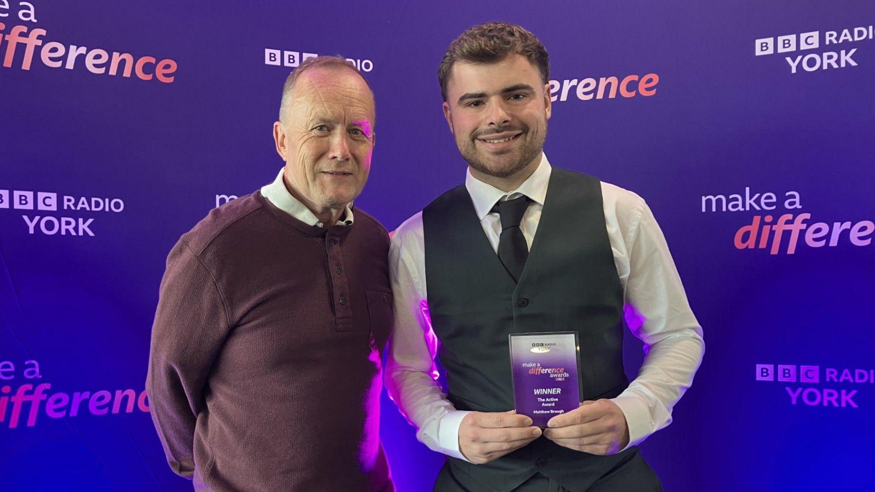 Two men are stood next to each other. On the right is a young smiling man with short brown hair, who is wearing a white shirt, grey waistcoat and dark grey tie. He is holding a glass award that says 'BBC Make A Difference Awards 2025 - Winner'. Next to him is an older man wearing a burgundy jumper over a white shirt. He is also smiling. Behind them is a backdrop that says 'BBC Radio York' and also 'BBC Make A Difference'. 