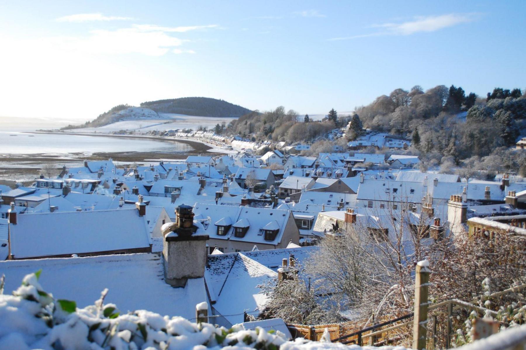 The picture has been taken from a vantage point looking out over the rooftops. The houses are flanked by the sea and a wooded hillside. The sun is shining.