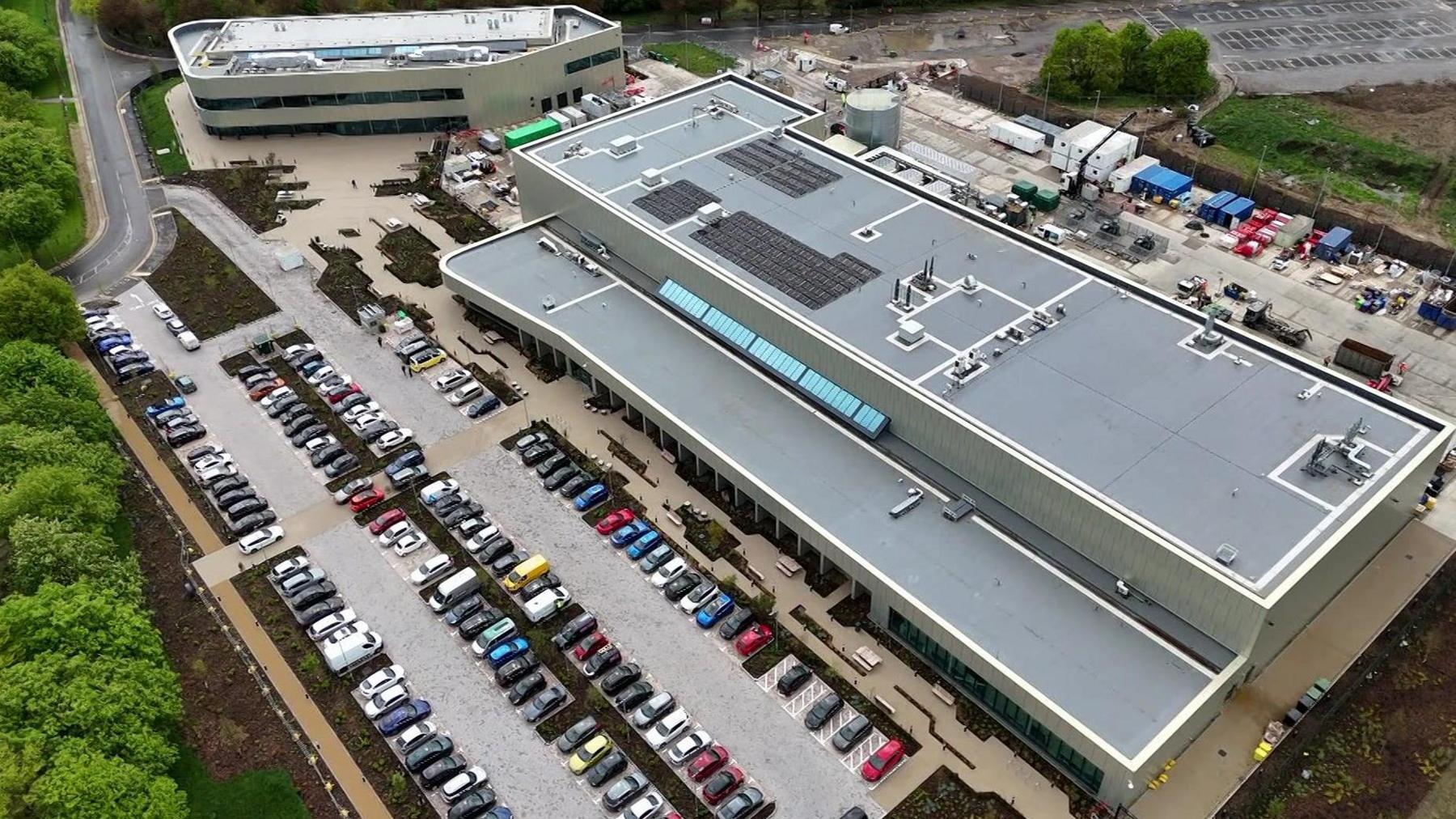 A drone shot of a large, long, modern-looking building, and a smaller one next to it. There is a car park with about 100 cars in it.