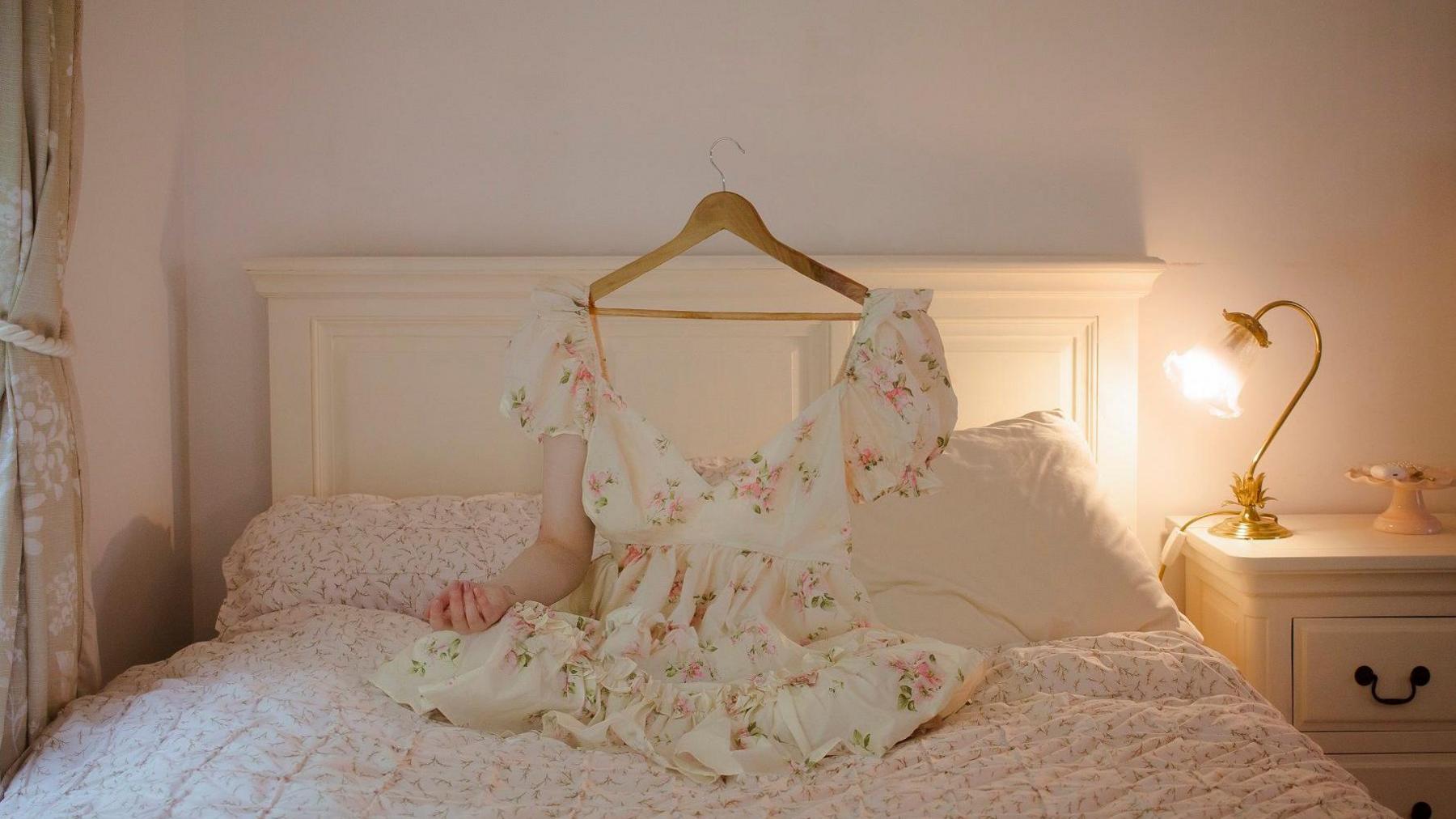 An artwork showing a woman sitting on a bed wearing a white dress. However, one arm is invisible, as is her head, which is replaced by a coat hanger. The image represents the way some disabled people feel invisible.