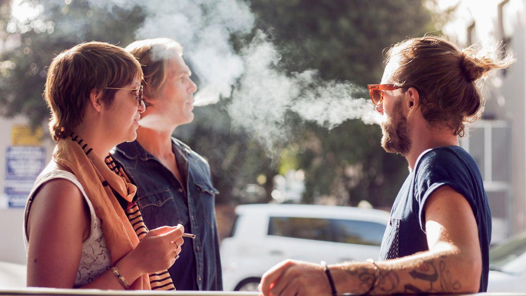 Three people are standing together outdoors - one woman has a cigarette in her hand while the other two men are blowing tobacco smoke out of their mouths