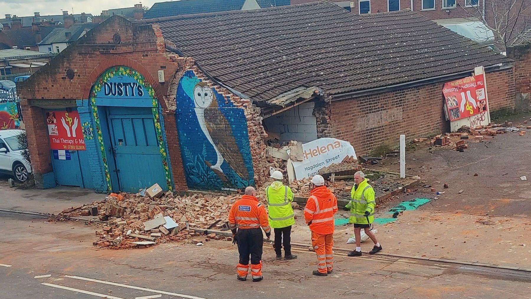 Old building with section of brick wall demolished. There is a pile of bricks in front of the wall and four people - wearing construction-type clothing are standing next to the building. A picture of an owl is painted on the building front wall and the name Dusty's is written above the front doors.