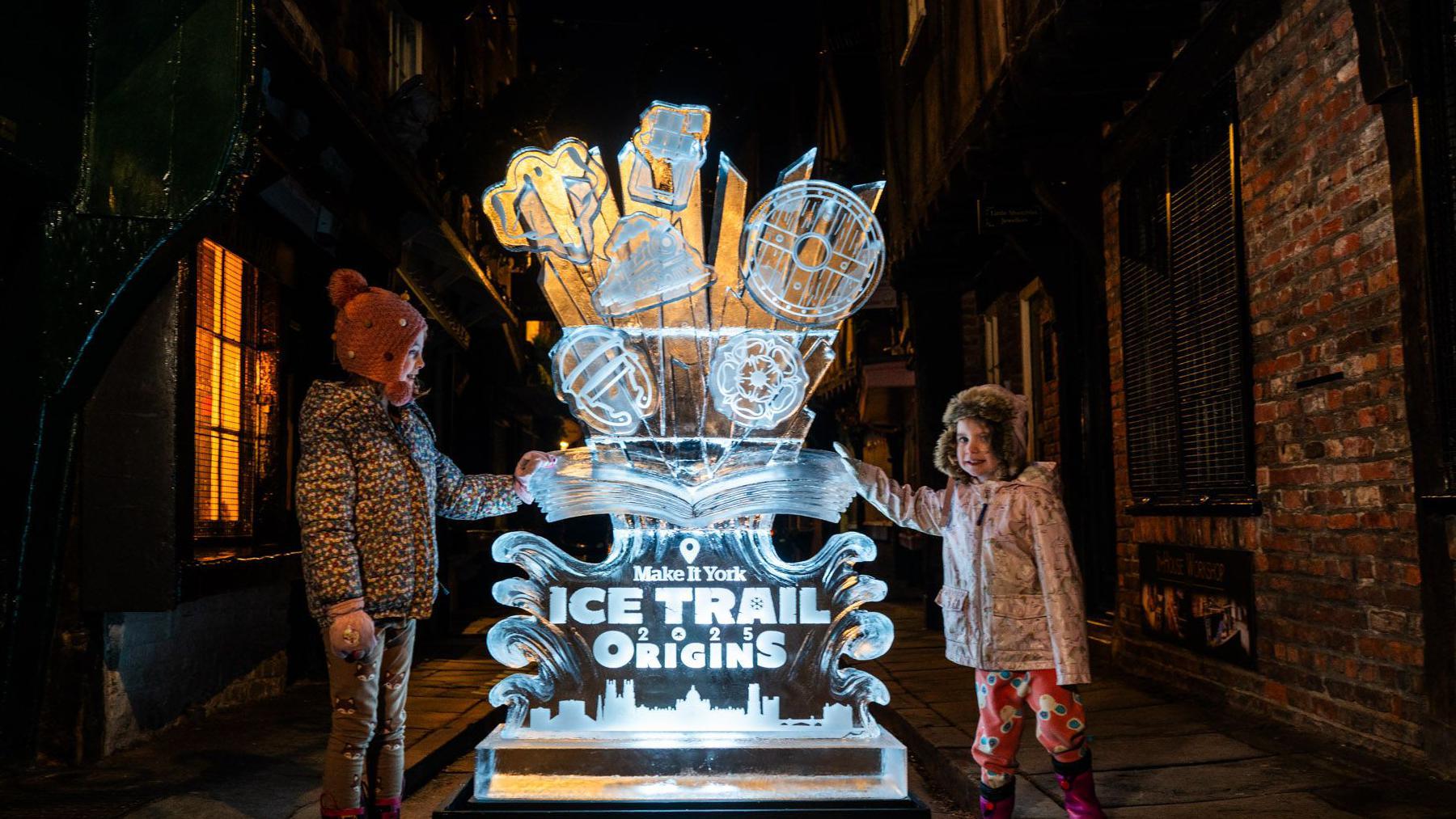 An ice sculpture in the centre of the image, with two children either side of it both wearing coats on a street in York. With the words Make it York, Ice Trail 2025 Origins carved on the sculpture