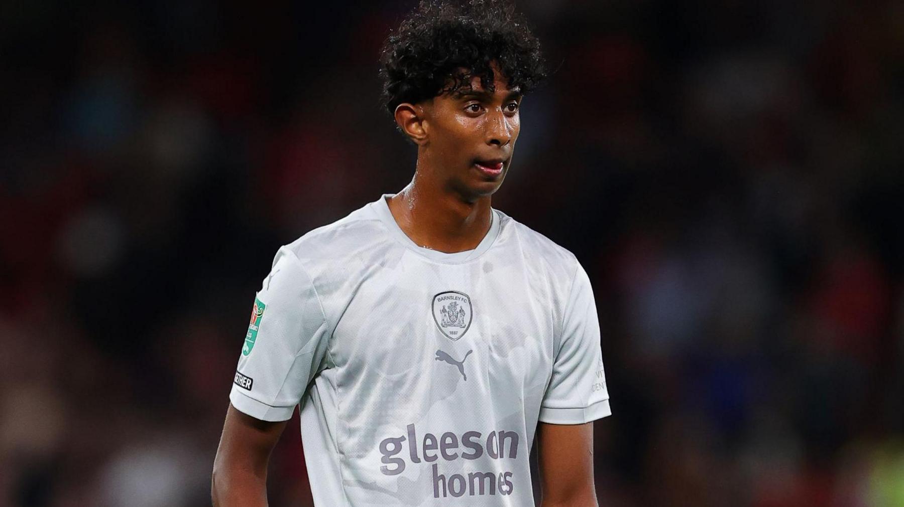 Vimal Yoganathan on the pitch in his pearl-grey Barnsley kit. Vimal, sweating, looks focused on the game and a crowd is blurred behind him. 