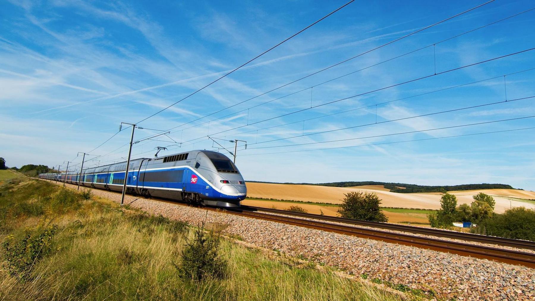 A high speed SCNF train in France passing through a field. There is a blue sky with very wispy clouds overhead, some rough grass on the left of the track and yellow fields on the right side which look farmed. The train is silver and blue with the red SCNF logo on