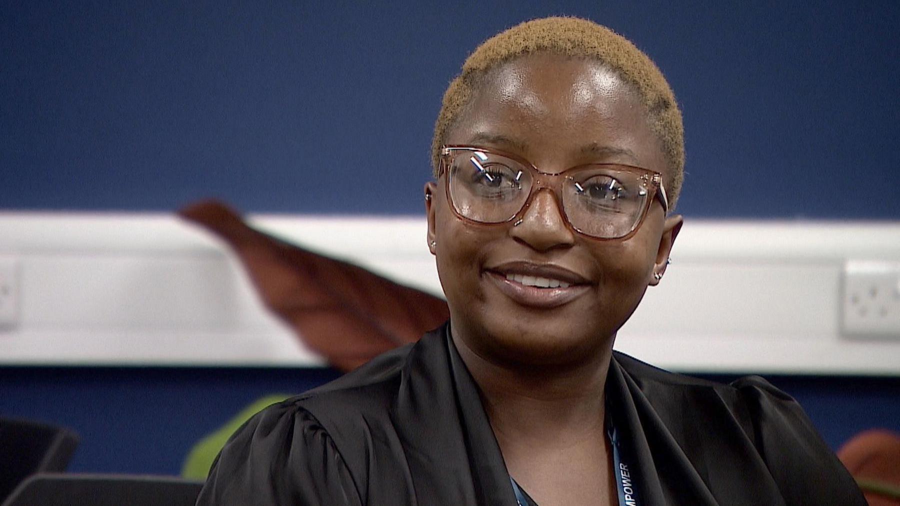 Natalie, a woman wearing glasses an a black top is smiling as she looks slightly to the left of the shot. She is indoors, in a room with dark blue walls and a white panel with plug sockets.