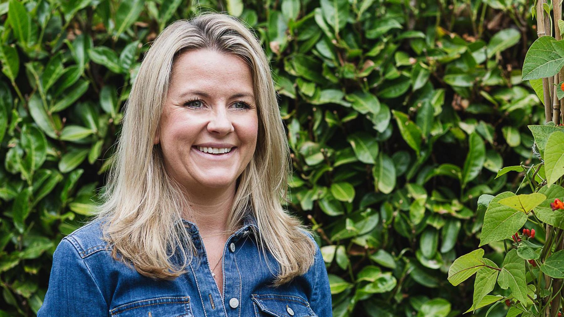 A woman is shown from the chest up, smiling and looking away from the camera. She has shoulder-length blonde hair and is wearing a blue denim jacket top. She is stood in front of a leafy green hedge.