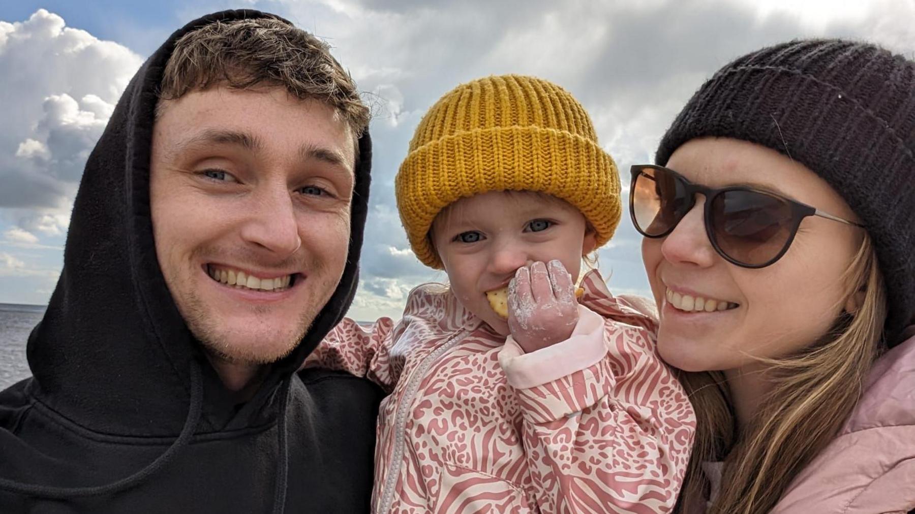 A man, woman and young child taking a selfie on a beach while the child eats a cracker.