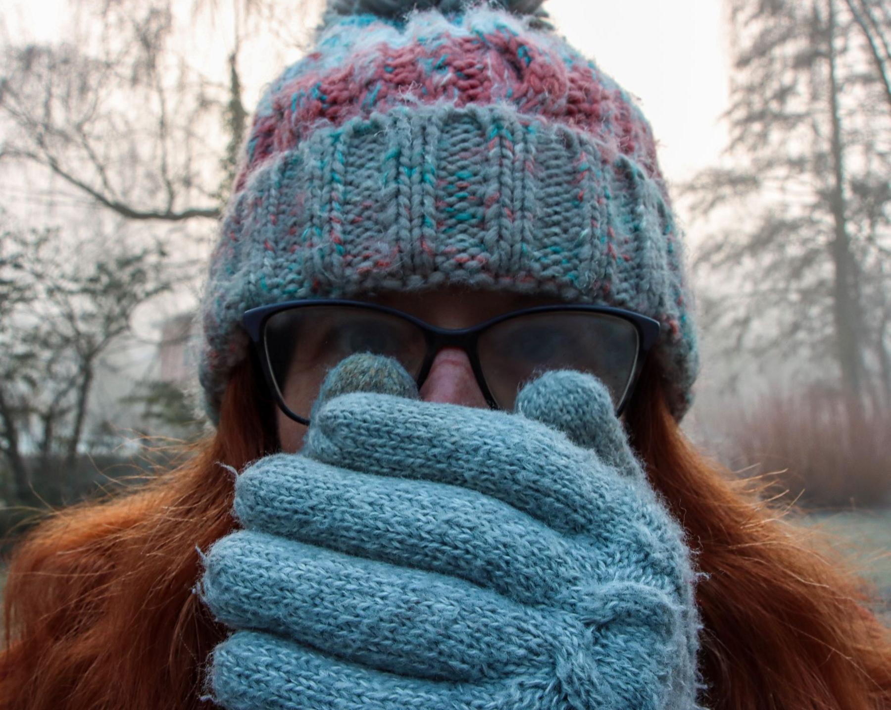 Close-up picture of a woman wearing blue woollen gloves. The woman, who has long red hair and a blue woollen hat on, is holding her hands over her face, as if trying to warm them with her breath. She is wearing glasses and is looking directly at the camera.