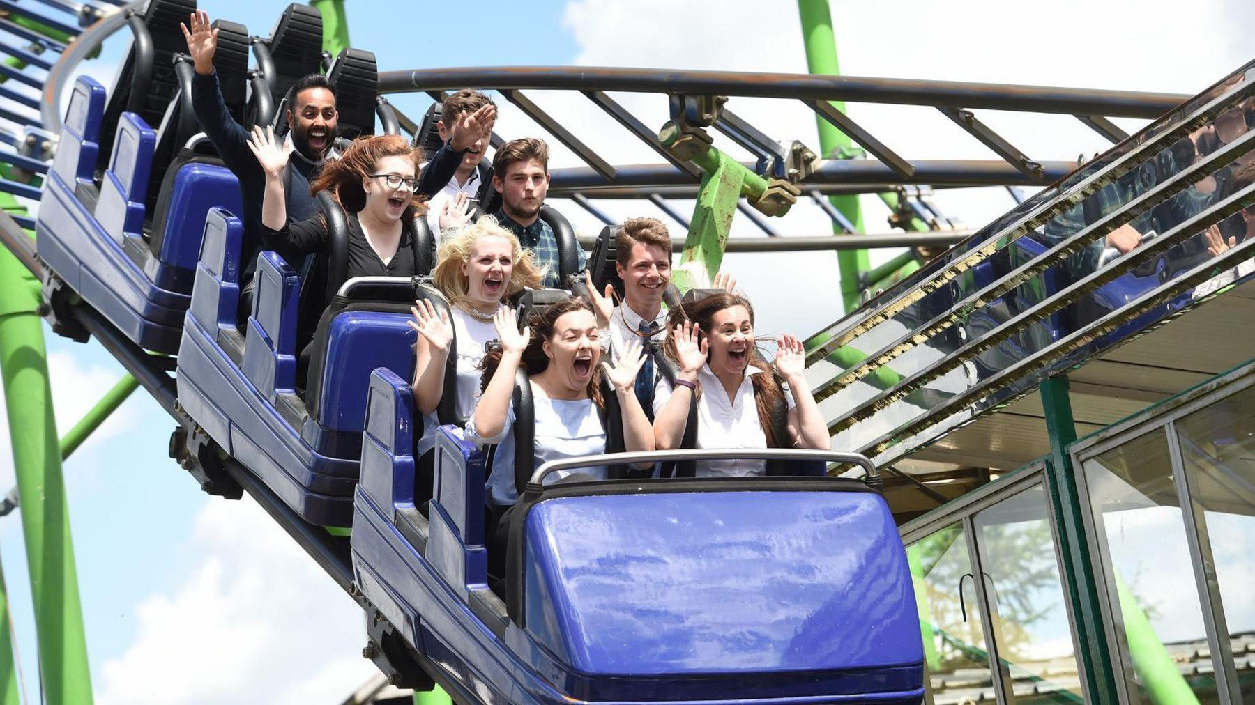 Blue rollercoaster cars on a rollercoaster track with people holding up their hands as the cars negotiate a steep downhill section. A catwalk is visible to the right of the ride.