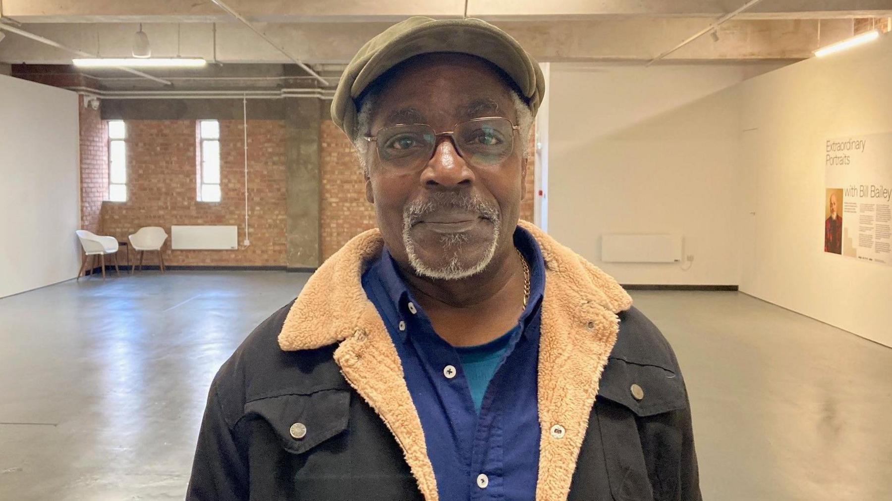 A man wearing a flat cap and glasses and fur-lined coat standing in an empty art gallery