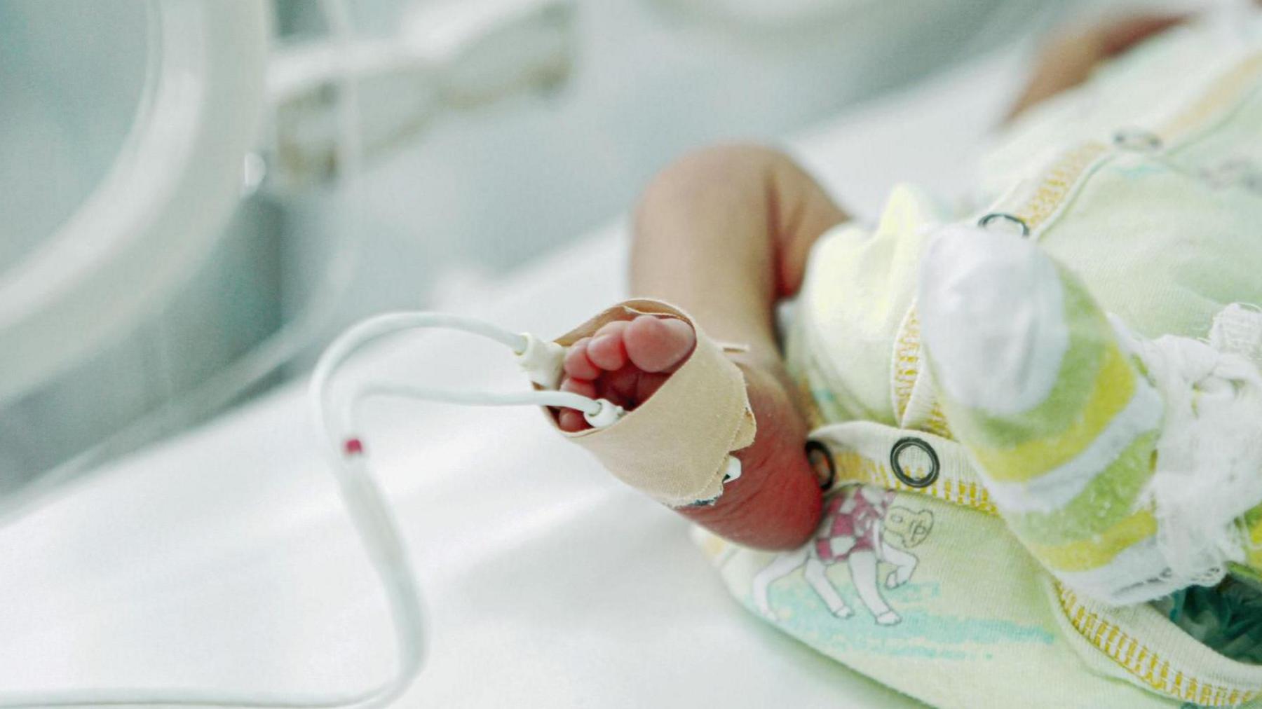A stock image of a premature baby lying in an incubator in hospital, wearing a baby grow. It is a close up of the baby's legs with medical tubes coming from its foot.