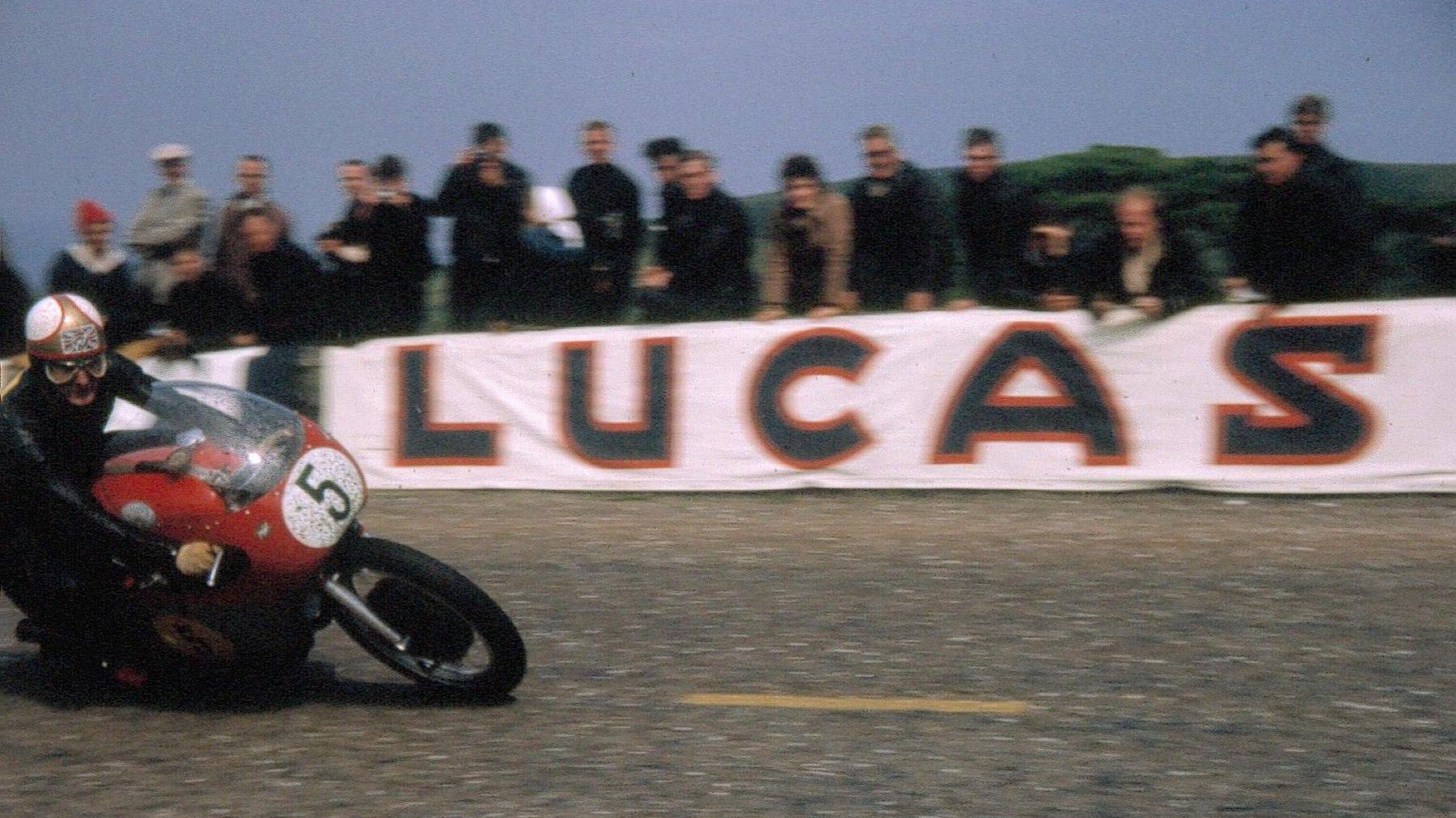 Mike Hailwood on a red bike with the number 5 he waears a white helmet and goggles, onlookers watch and there is a banner that reads Lucas.