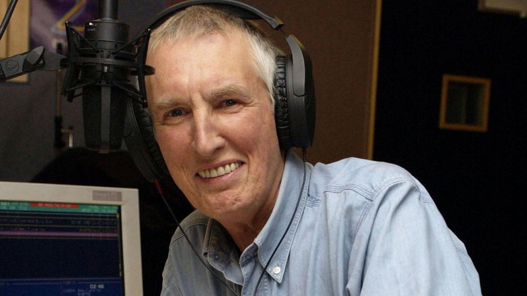 Johnnie Walker, wearing headphones, next to a microphone in a radio studio. He has short white and brown hair and wears a button down collared chambray shirt. There is an older style computer in the background. The image is from 2004. 