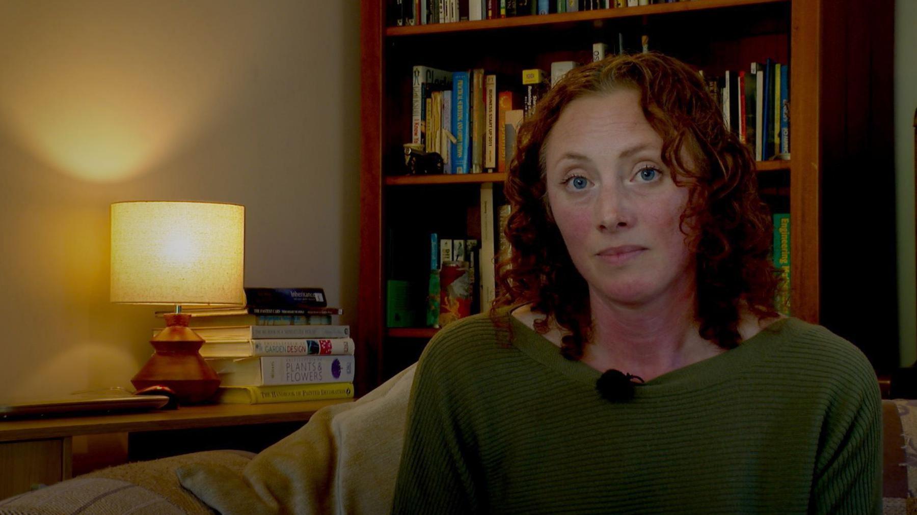 Naomi Keegan with red curly hair and green woollen jumper, looking at the camera with a serious face, with a lamp on a table and a bookshelf full of books behind her