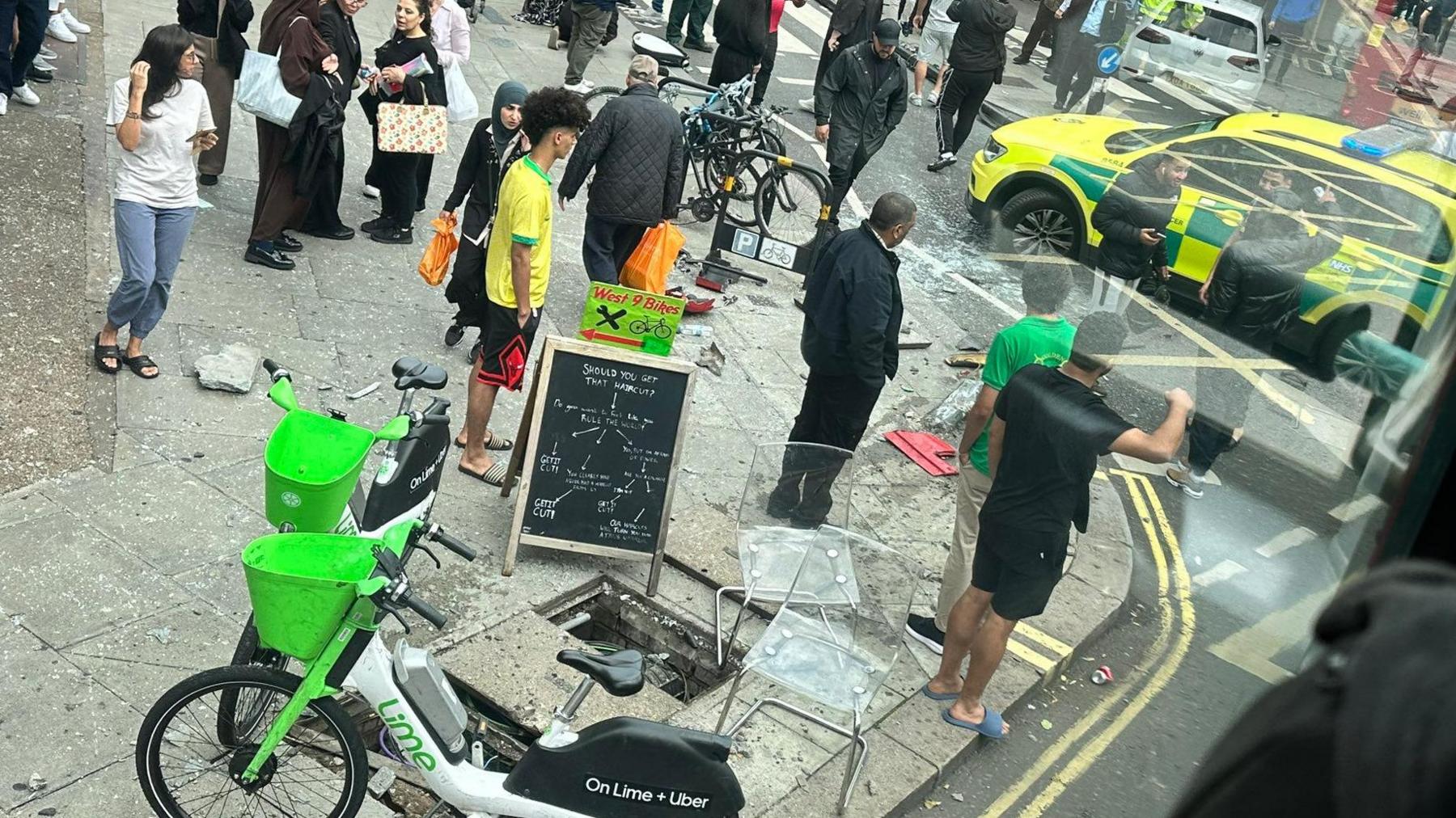 Image taken from what appears to be upper deck of a bus shows people gathered on a street corner, some watching while others walk by, there are Lime rental bikes in the foreground and an emergency vehicle in the road in the background