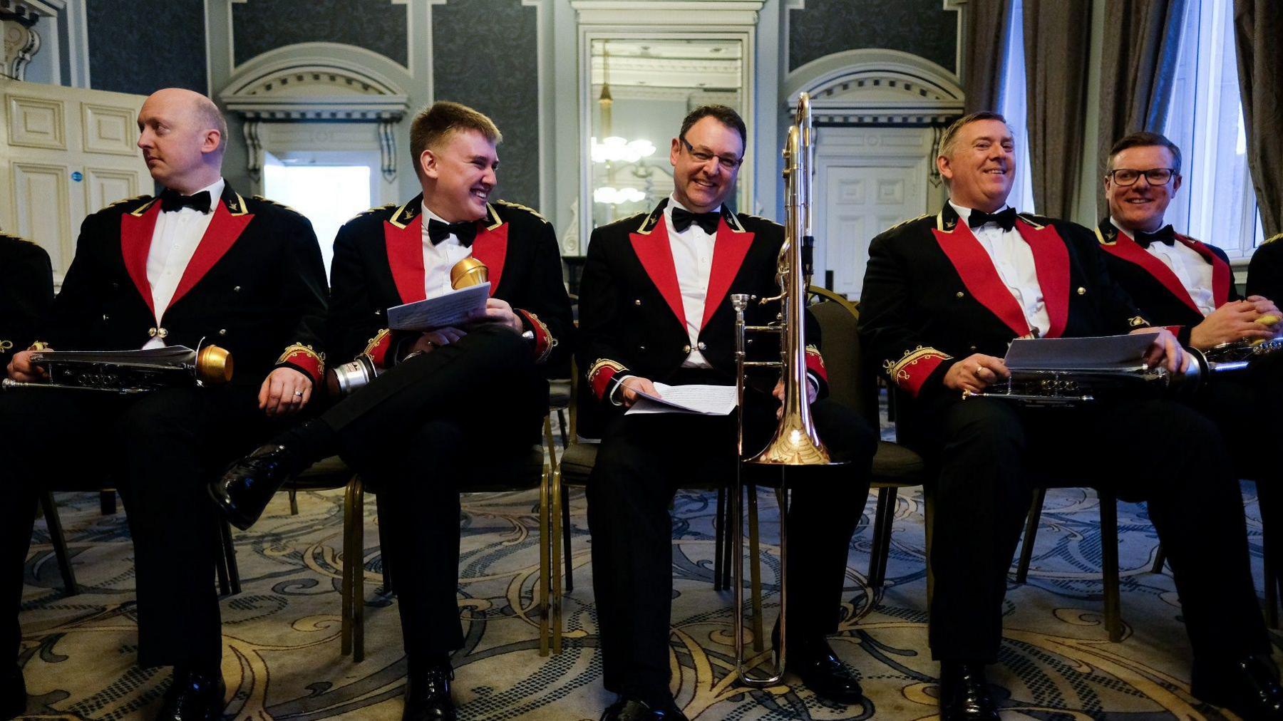 Members of Black Dyke Brass Band wait to perform at Huddersfield Town Hall. They are sitting on chairs in black, red and gold uniforms holding their music and instruments.