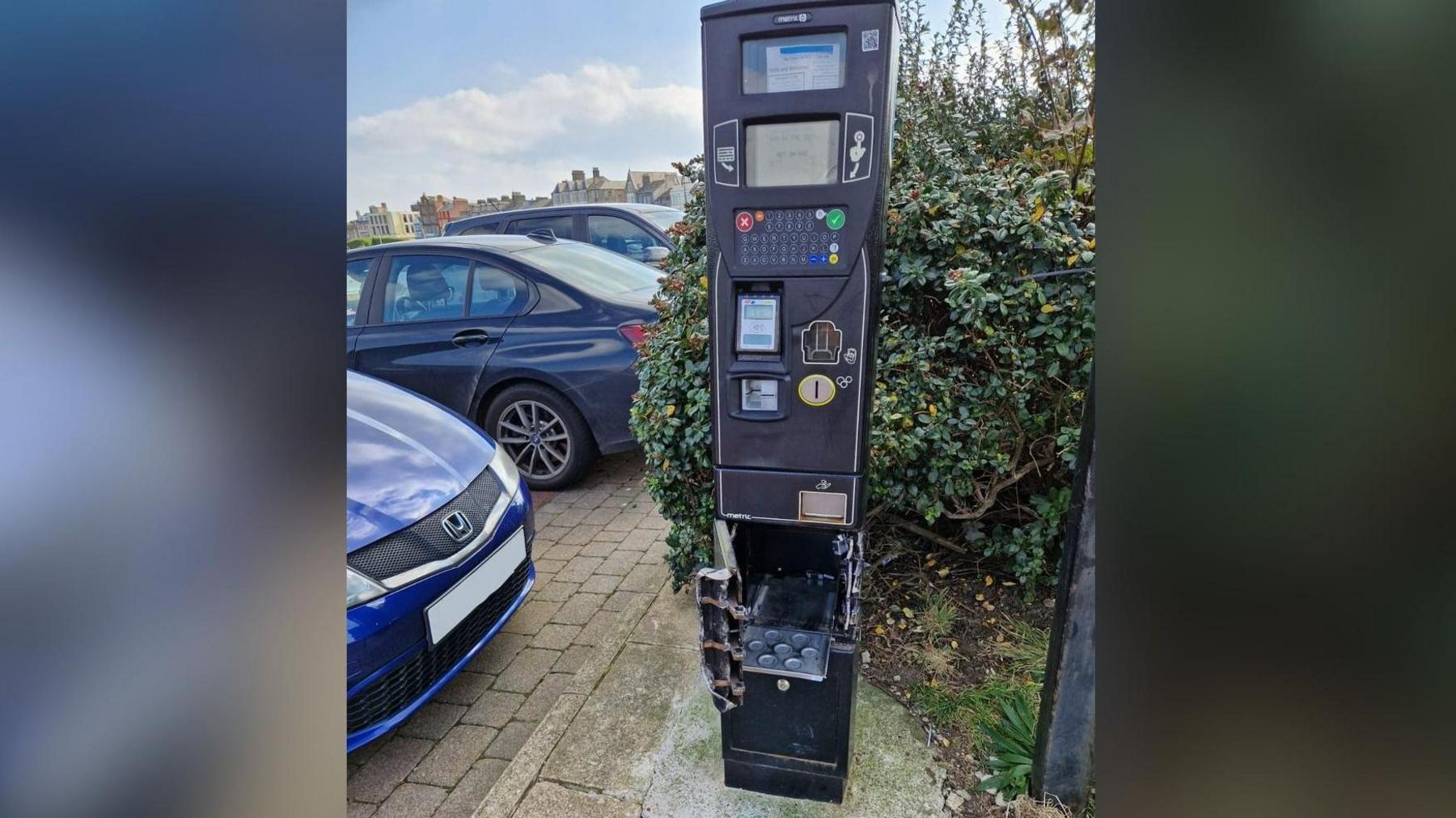 A pay and display machine showing damage to the bottom section where the cash box would have been, with the broken door left open. Some cars can be seen parked next to it.