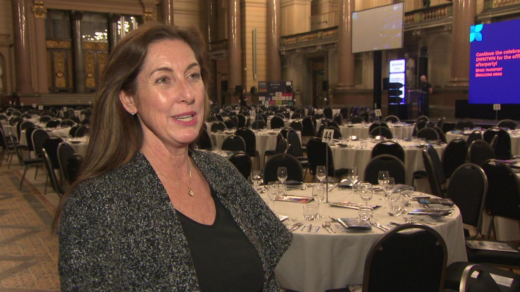 Woman with long brown hair wearing black top and grey cardigan in large hall with dining tables set out for an event