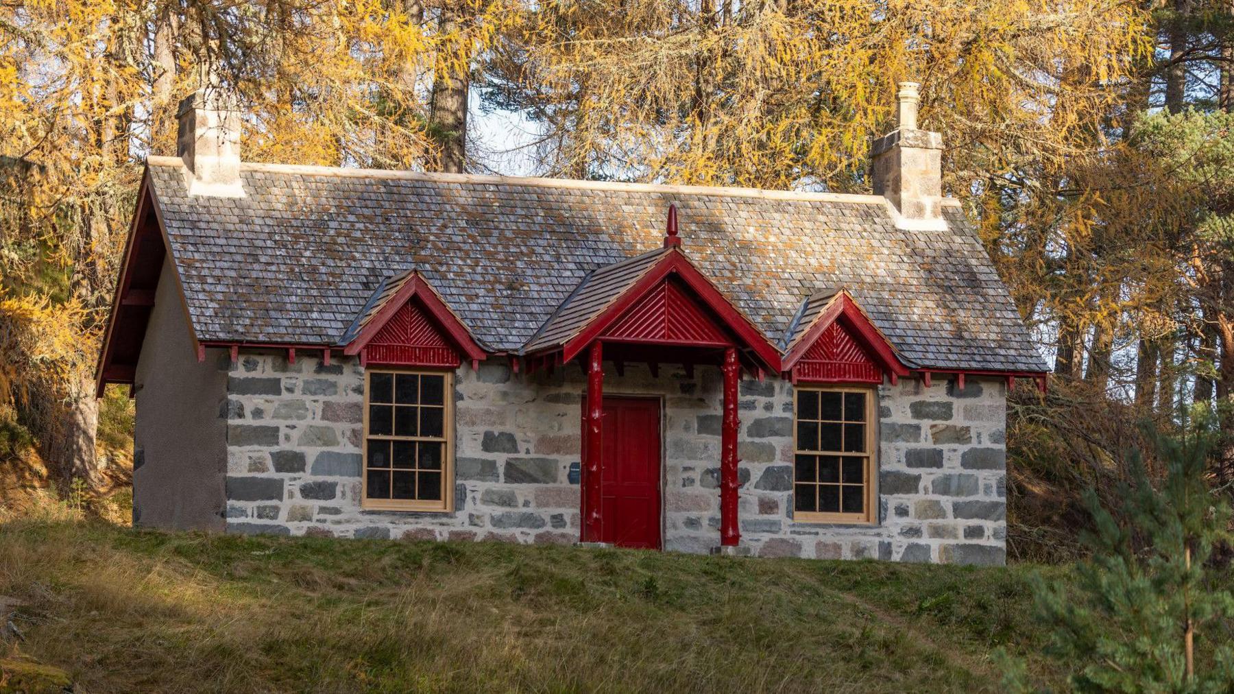 A 19th century cottage in the woodlands. The door is red with a red porch over the door. There are two windows on either side of the red door in the middle. Two chimneys are on either side of the roof. 