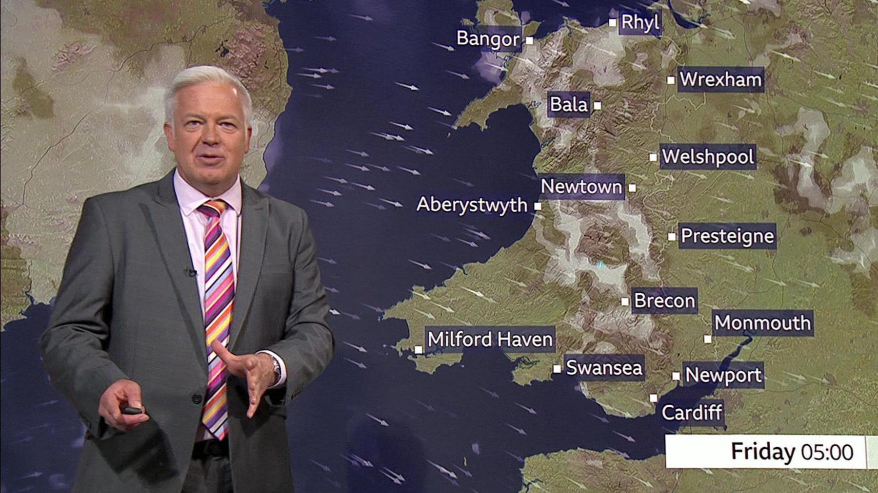 Derek Brockway wearing a grey suit, a colourful tie and pink shirt. She is standing in front of a weather map showing Wales and various towns and cities. 