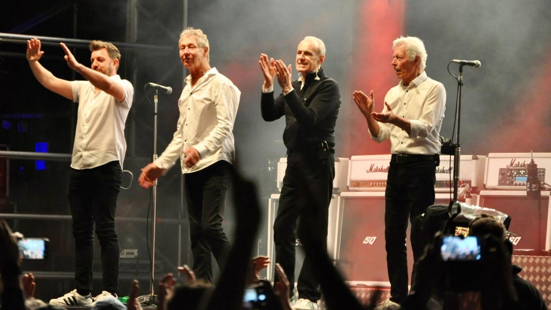 Four members of the band standing on stage thanking the audience at the end of their performance. They are all wearing black trousers and a white shirt, with Francis Rossi also wearing a black zip up jumper. They are clapping and smiling at the audience