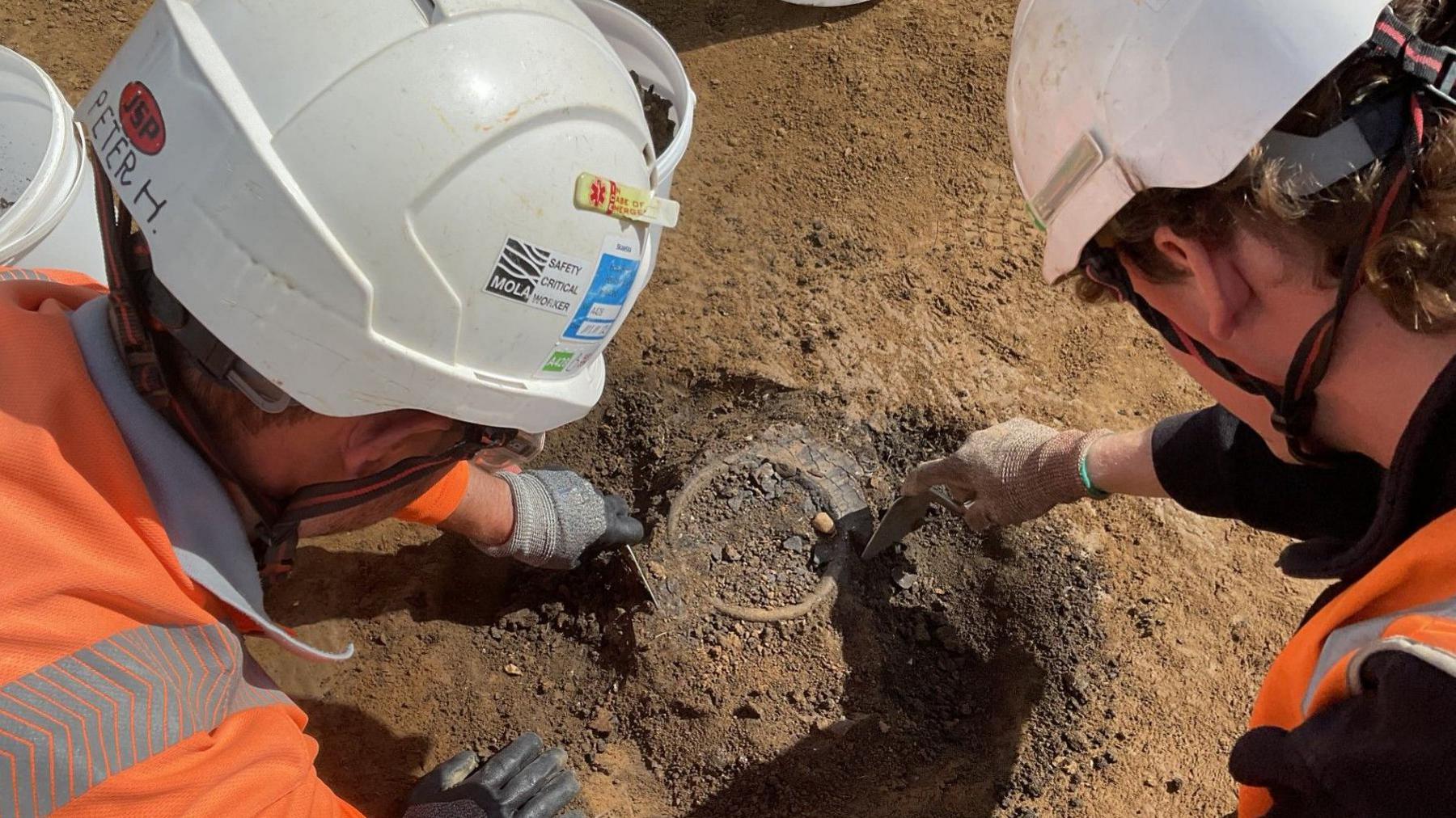 There are two people wearing white helmets and orange high vis vests. Both people are looking at the ground as they are holding metal tools and scraping earth from around a circular artefact.