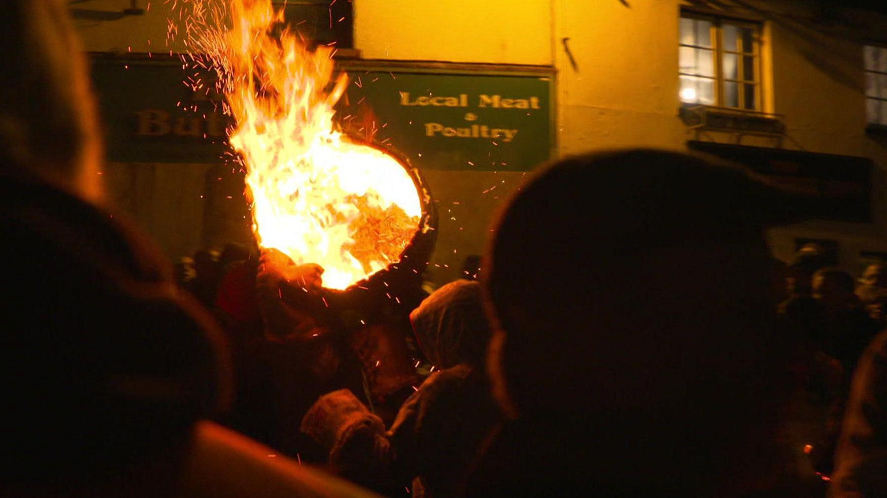 A flaming barrel is carried through the air past a local meat and poultry shop. It is dark and flames are shooting from the barrel.