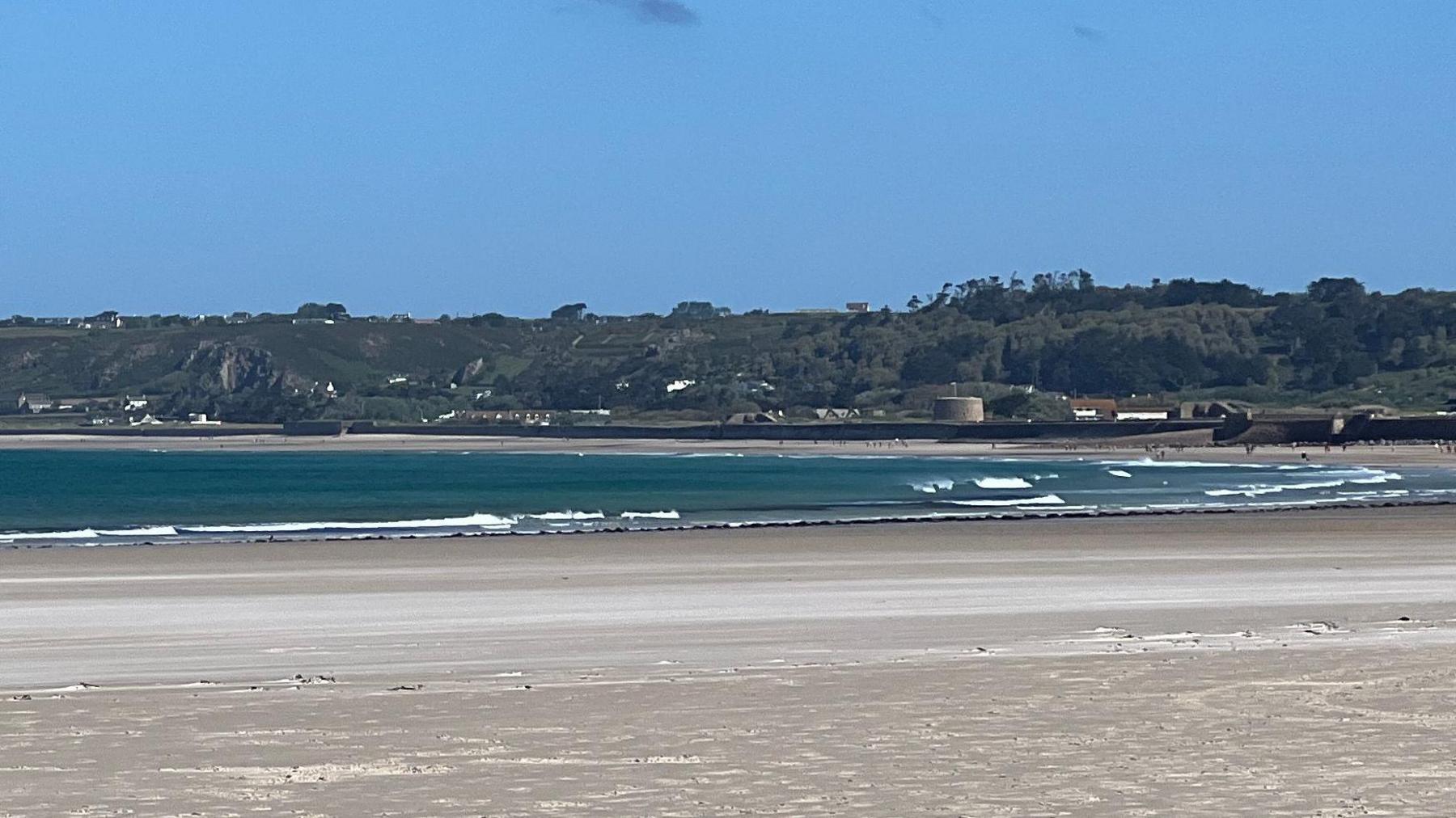 A picture of St Ouen in Jersey. It is a sandy beach and in the distance there is blue water. In the distance there is green land with buildings along the shore.