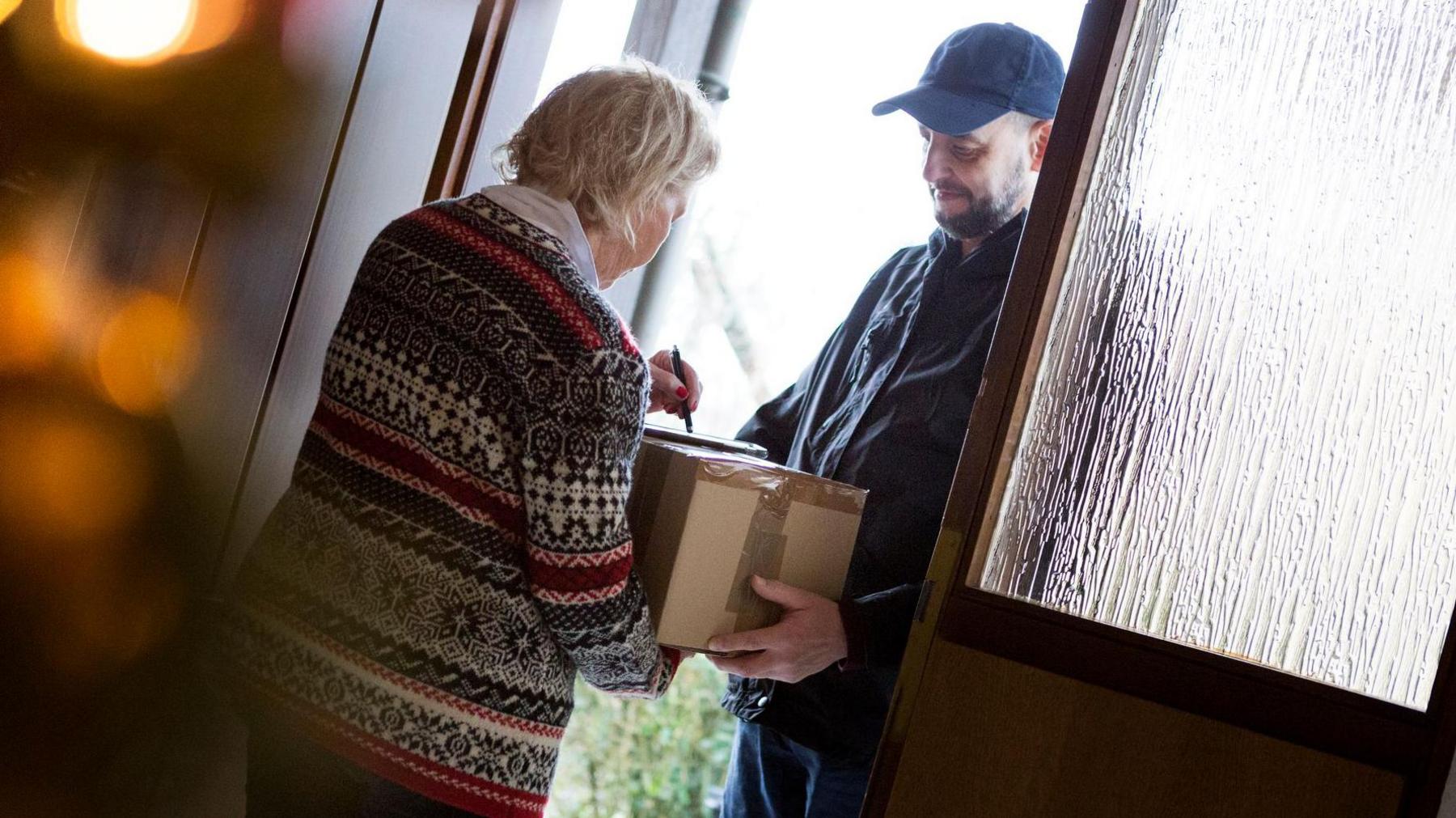 A woman in a festive jumper that is blue, red and white is signing for a parcel at a door. A deliveryman, dressed in a black coat and baseball cap, is holding the parcel.