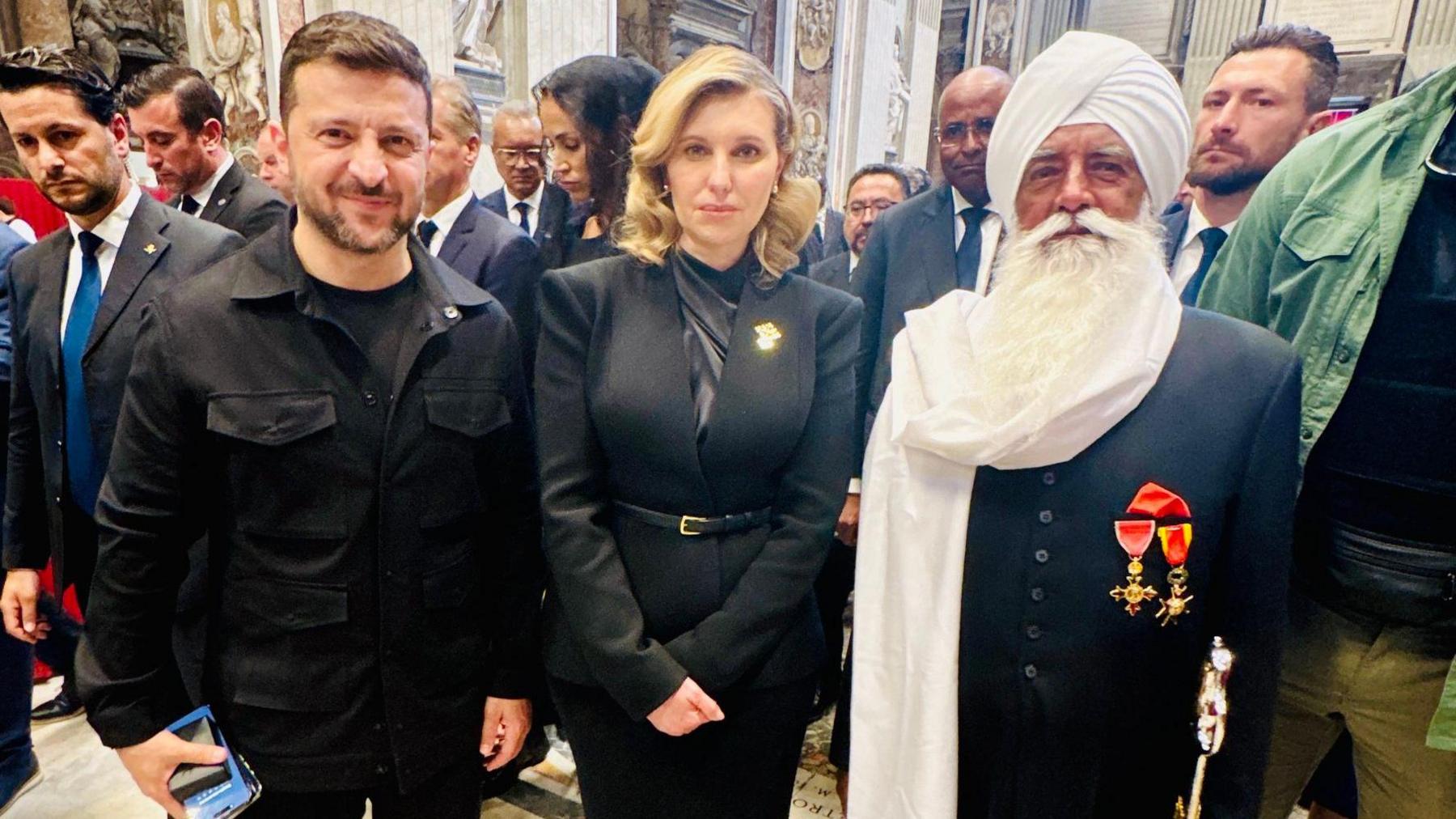 The Ukrainian president, Volodymyr Zelensky, a man with short dark hair, is wearing a black T-shirt under a black shirt. He is standing next to his wife, Olena Zelenska, who has shoulder-length blonde hair and is wearing a black suit. They are with a bearded man, Bhai Sahib, Professor Mohinder Singh, who is wearing a long black jacket two medals.