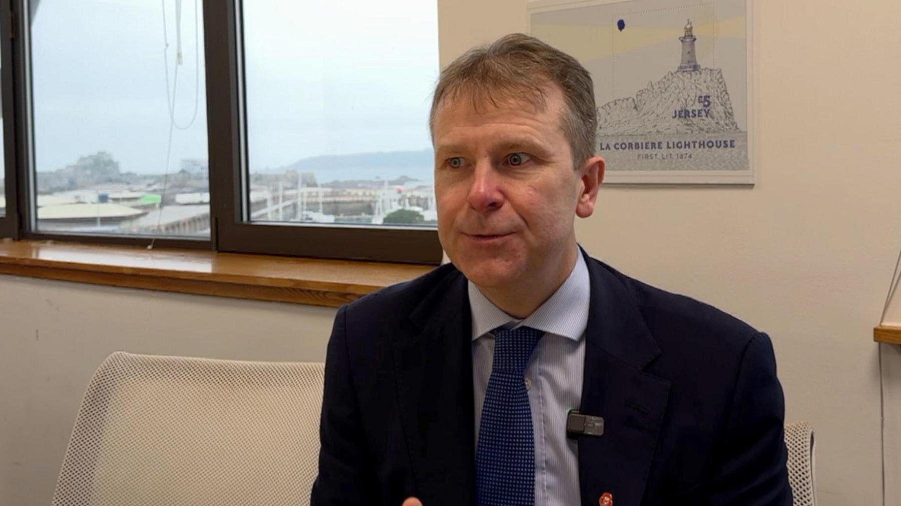 Matt Thomas, a man wearing a dark blue suit, light blue shirt and royal blue tie. He has short brown hair. He is sitting in front of a print of La Corbiere Lighthouse, and a window through which the marina is visible.